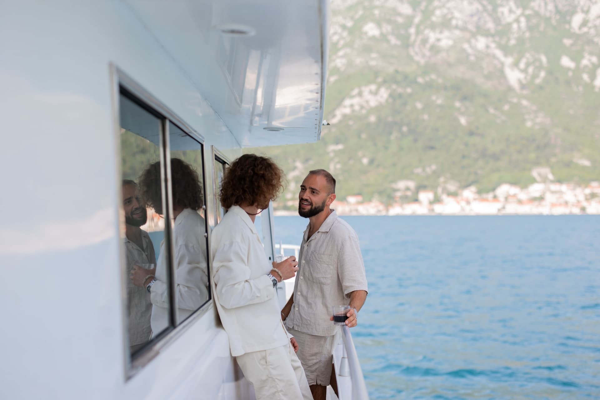 Two guests talking on the side deck of Marea as the shore of the bay passes behind them
