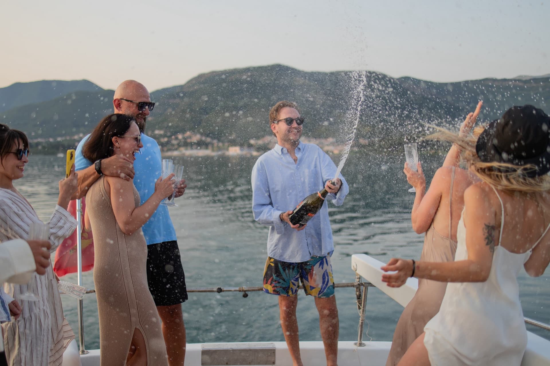 Guests on deck with champagne being opened, the mountains of the bay behind them